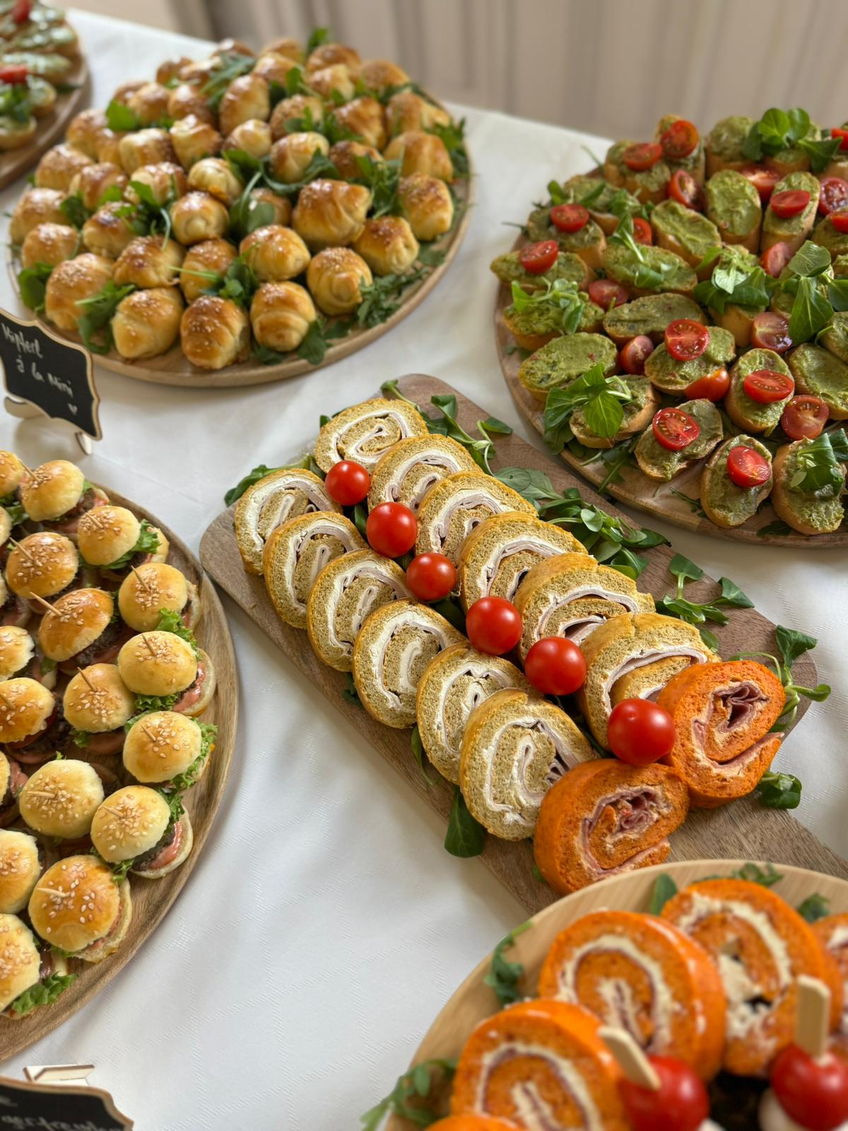 Holzplatten mit gerollten Wraps, Mini-Burgern, Tomatenhäppchen und frischen Kräutern auf weißem Tischtuch.