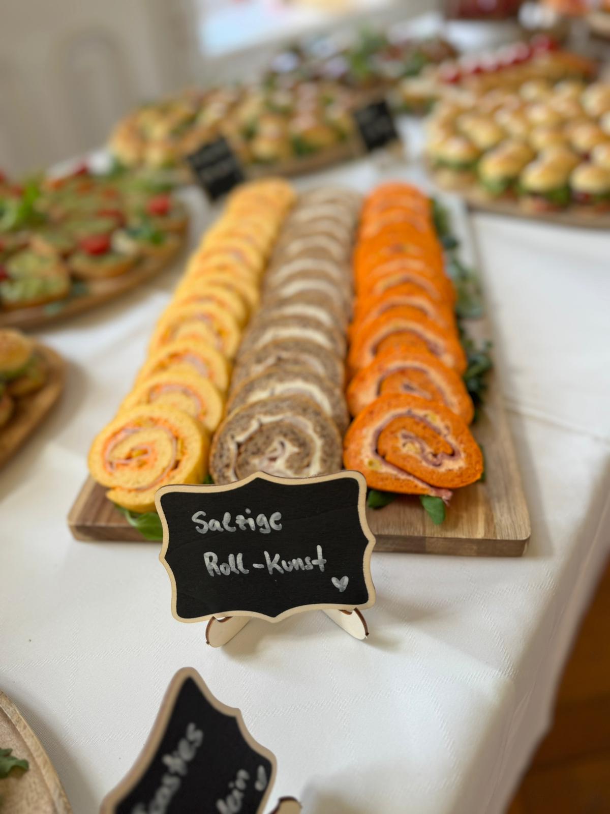 Holzplatte mit drei Reihen gerollter Wraps in verschiedenen Farben und Füllungen, dekoriert mit Rucola, auf weißem Tischtuch.