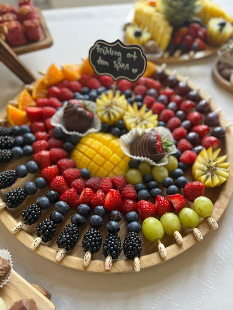 Runde Holzplatte mit frischen Früchten, dekorativ angerichtet in Kreisen, mit Schild „Frühling auf dem Spieß“.