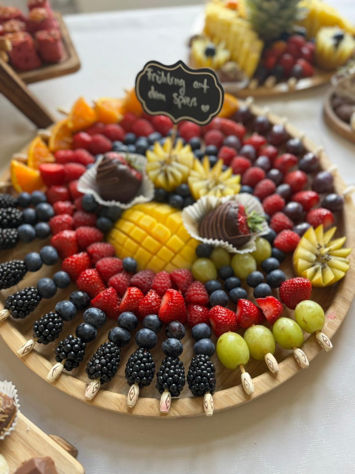 Runde Holzplatte mit frischen Früchten, dekorativ angerichtet in Kreisen, mit Schild „Frühling auf dem Spieß“.